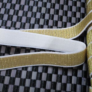 Puede incluir: Borde elástico blanco con purpurina dorada. El borde está enrollado y muestra el respaldo blanco.