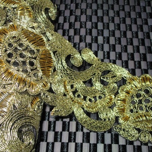 May include: Close-up of gold metallic lace trim with intricate floral and swirl designs. The trim is set against a black and white checkered background, highlighting the detailed embroidery and texture of the lace.