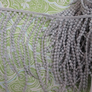 May include: Grey pom-pom trim with a beaded fringe. The trim is made of soft, fluffy yarn and has a subtle sheen.