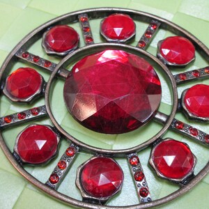 Könnte beinhalten: Ein runder, metallener Anhänger mit einem großen, roten, facettierten Edelstein in der Mitte. Der Anhänger ist von einem Ring aus kleineren, roten, facettierten Edelsteinen umgeben.