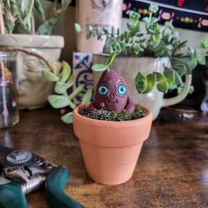 Puede incluir: Una pequeña maceta de barro marrón con la parte superior de musgo verde. Una figura de barro marrón con un brote y ojos azules está sentada en la maceta.