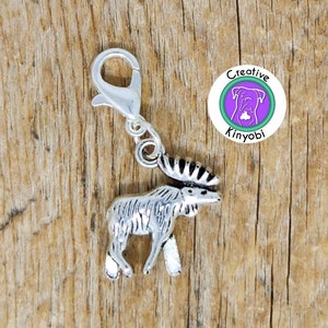 Puede incluir: Encanto de alce plateado con un clip en la parte superior. El encanto está sobre una superficie de madera. El encanto es parte de un conjunto de encantos de animales. El encanto es para un tirador de cremallera o un encanto de bolso.