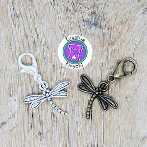 May include: Two silver and bronze dragonfly charms with a lobster clasp. The charms are on a wooden background.