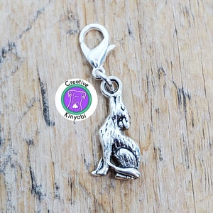 Puede incluir: Encanto de plata con un diseño de lobo sentado. El encanto tiene un cierre de langosta de plata y está unido a un círculo morado y verde con el texto "Creative Kinyobi".