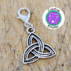 Puede incluir: Encanto de metal plateado con un diseño de nudo celta. El encanto tiene un cierre de langosta para sujetarlo a una bolsa, llavero u otros artículos. El encanto está sobre una superficie de madera.