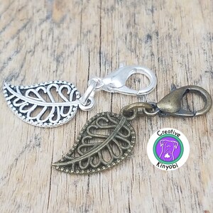 May include: Two silver and bronze leaf-shaped charms with a silver and bronze lobster clasp. The charms are attached to a silver and bronze chain. The charms are on a wooden surface. The charms have a decorative design. The charms are labeled "Creative Kinyobi".