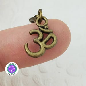 Puede incluir: Un pequeño dije con el símbolo Om de color bronce. El dije es un colgante con un lazo en la parte superior para sujetarlo a un collar o pulsera. El símbolo Om es un icono espiritual en el hinduismo y el budismo.
