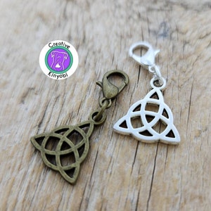 May include: Two silver-toned and bronze-toned metal Celtic knot charms with a lobster clasp. The charms are in the shape of a triangle with a knot design. The charms are on a wooden surface.