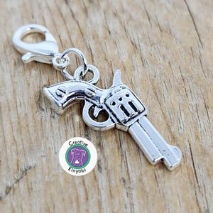 May include: Silver-colored revolver charm with a lobster clasp. The gun charm is detailed with a trigger, hammer, and cylinder. The charm is attached to a silver clasp, ready to be added to a bracelet or necklace.