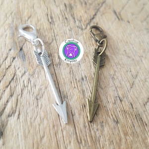 May include: Two silver and bronze arrow charms with a clip on the top. The charms are on a wooden surface.
