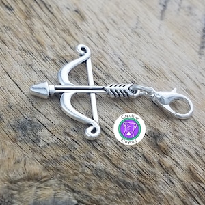 Può includere: Ciondolo in argento a forma di arco e freccia con un fermaglio in argento. Il ciondolo è attaccato a una superficie in legno. Il ciondolo ha un piccolo cerchio bianco con il testo "Creative Kinyobi" in nero.