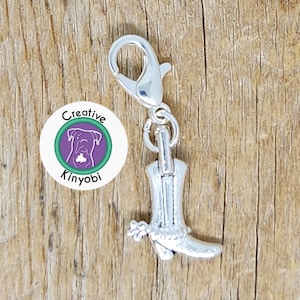Puede incluir: Un charm plateado con forma de bota de vaquero con detalles florales, sujeto a un cierre de mosquetón. El charm está junto a un logotipo redondo con el texto "Creative Kinyobi" y una ilustración de perro.