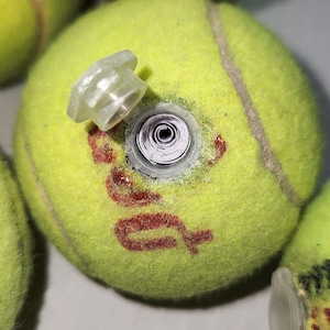 May include: A close-up of several yellow tennis balls, one with a clear plastic insert and a red logo. The insert is partially removed, revealing a rolled-up paper. The tennis balls are on a white surface.