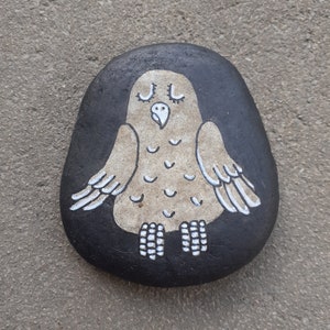 Puede incluir: Una piedra pintada a mano con un diseño de búho blanco sobre un fondo negro. El búho tiene los ojos cerrados y un pequeño pico.