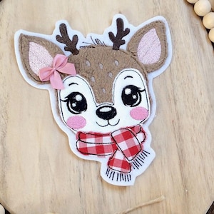 May include: Embroidered applique of a brown deer with pink ears and a red and white checkered scarf. The applique is on a wooden background with a brown woven border.