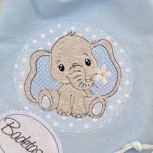 Könnte beinhalten: Eine hellblaue Kordeltasche mit weißem Punkte-Muster. Die Tasche hat ein gesticktes Design eines grauen Elefanten, der eine weiße Blume hält. Die Tasche hat ein weißes ovales Etikett mit der Aufschrift "Badetasche" in Schwarz.