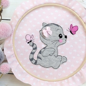 Könnte beinhalten: Ein rosa-weißer bestickter Reifen mit einem grauen Kätzchen, das eine rosa Schleife und einen Schmetterling trägt. Der Reifen ist mit kleinen rosa Herzen verziert.