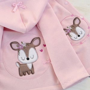 Può includere: Una giacca rosa con cappuccio con un cervo marrone ricamato sul davanti e un cervo più piccolo sulla tasca posteriore. La giacca ha un fiocco sul cappuccio e si chiude con bottoni a pressione sul davanti.