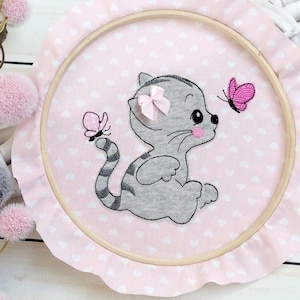 Könnte beinhalten: Ein rosa-weißer bestickter Reifen mit einem grauen Kätzchen-Design. Das Kätzchen trägt eine rosa Schleife und hat einen Schmetterling daneben.