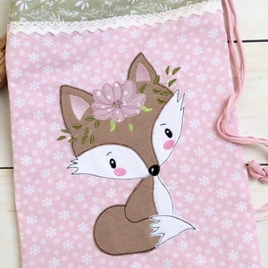 May include: A pink drawstring bag with a white lace trim and a brown fox with a pink flower crown embroidered on the front.