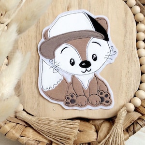 May include: A brown and white felt fox applique wearing a white felt hat with a brown brim. The applique is sitting on a wooden surface with a woven border.