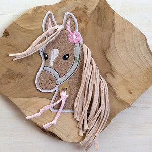 Puede incluir: Una aplicación de cabeza de caballo marrón con un detalle de flor rosa y cinta rosa. La aplicación está sobre una superficie de madera.