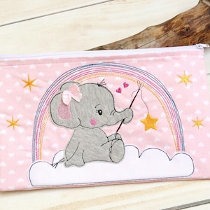 Puede incluir: Una bolsa rosa con cremallera y ribete blanco. La bolsa presenta un diseño bordado de un elefante gris que sostiene una caña de pescar y una estrella. El elefante está sentado en una nube blanca con un arcoíris detrás. El fondo es una tela estampada rosa y blanca.