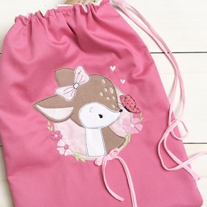 May include: Pink drawstring bag with a cute embroidered fawn design. The fawn is brown with white spots and has a pink bow on its head. The fawn is surrounded by pink flowers and a butterfly.