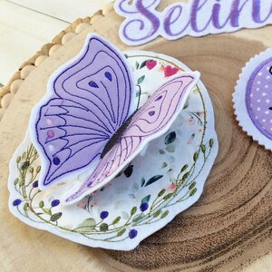 May include: A wooden slice with embroidered patches. A purple butterfly patch with a floral design, a name patch that says "Selina", and a round patch that says "Schulkin 2025".
