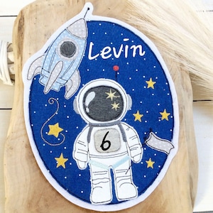 Puede incluir: Una aplicación de fieltro azul y blanco con un cohete, un astronauta y estrellas. La aplicación dice "Levin" y "6" en texto blanco.