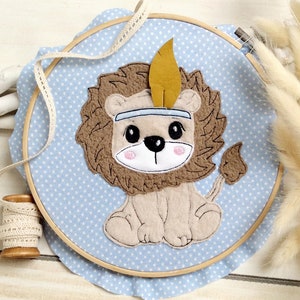 May include: A brown and beige felt lion with a yellow feather headdress on a blue and white polka dot fabric background. The lion is sitting with its paws together and has a cute, cartoon-like face.
