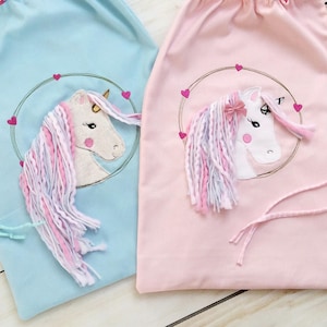 Puede incluir: Dos bolsas con cordón, una celeste y otra rosa, cada una con un diseño de unicornio bordado. Los unicornios tienen caras blancas, crines de hilo rosa y morado, y están rodeados por una corona de corazones. Las bolsas son adecuadas para llevar objetos.