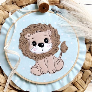 May include: A light blue hoop with a white lace trim and a brown embroidered lion with a fluffy mane. The hoop is on a woven brown basket.