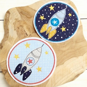 Puede incluir: Dos parches redondos de fieltro con cohetes y estrellas bordadas. Un parche tiene un fondo azul con estrellas blancas y un cohete amarillo. El otro parche tiene un fondo azul claro con lunares blancos y un cohete gris.
