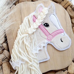 Può includere: Una testa di cavallo bianca con un morso rosa e una criniera intrecciata di filato bianco. La testa del cavallo è fissata su una superficie di legno.