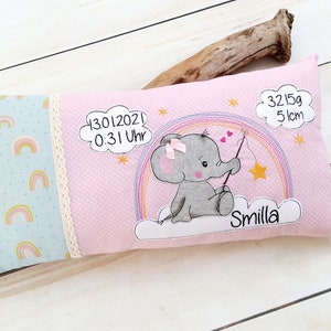 May include: A pink and blue baby pillow with a rainbow pattern and an embroidered elephant holding a star. The pillow also includes the text "13.01.2021 0:31 Uhr", "Smilla", "3215g 51cm".