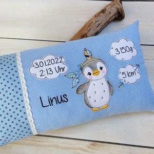 May include: A light blue baby pillow with a white polka dot pattern on one side. The other side features a cute cartoon penguin wearing a headband with feathers. The pillow also includes embroidered text: "30.12.2022 2:13 Uhr", "Linus", "3150g", and "51cm".