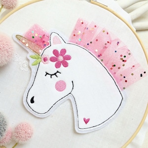 Könnte beinhalten: Ein weiß gesticktes Einhorn mit rosa Blumen und einem rosa Tüllrock. Das Einhorn hat ein geschlossenes Auge und ein rosa Herz an der Seite.