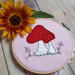 May include: A pink fabric hoop with a white and red mushroom embroidery. The mushroom has white polka dots on the red cap. The hoop is surrounded by a wooden frame.