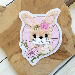 May include: A pink and brown embroidered bunny applique with a pink flower on its head. The bunny is surrounded by a pink circle with white polka dots.