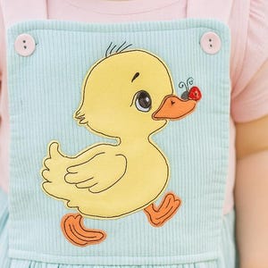 May include: A baby wearing a light blue corduroy overall dress with a yellow duck applique. The dress has a pink short-sleeved shirt underneath. The duck has orange feet and a red nose. The baby has blonde hair and is looking to the side.