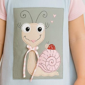 Peut inclure: Robe bleu clair avec manches et bordures roses. La robe présente un motif de colimaçon appliqué avec une coquille rose et rouge. L'escargot a un nœud rose et est sur un fond rectangulaire gris. La robe est portée par un jeune enfant.