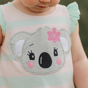 May include: A light pink and mint green striped dress with a koala bear applique. The koala has gray fur, pink cheeks, and a pink flower. The dress has ruffled sleeves and is designed for a toddler.