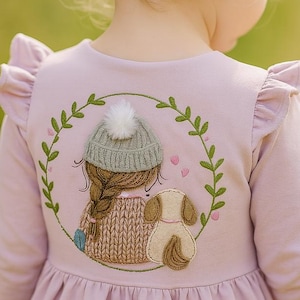 May include: A light purple long-sleeved dress with a decorative embroidered design on the back. The design features a girl with a braid, a dog, and a green wreath. The dress has ruffled sleeves and is worn by a young child.