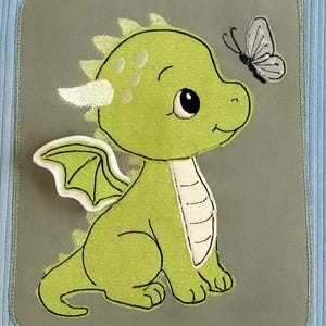 May include: Pale blue corduroy tote bag featuring a green dragon and butterfly appliqué. The dragon is depicted sitting with green wings and a white belly. The butterfly is grey and black.