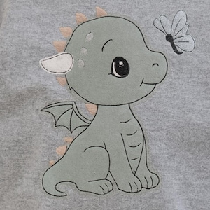 Könnte beinhalten: Ein graues Sweatshirt mit einem gestickten Cartoon-Drachen und einem Schmetterling. Der Drache ist hellgrün mit beigen Akzenten. Der Schmetterling ist weiß mit schwarzen Details. Das Sweatshirt hat einen Rundhalsausschnitt.