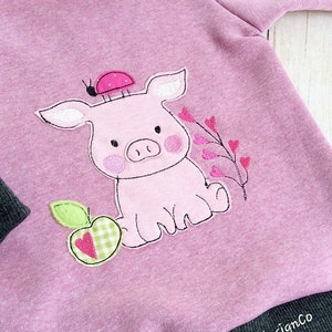 Op de afbeelding: Een roze longsleeve met een zwarte en grijze boord. De trui heeft een geborduurd ontwerp van een roze varken met een lieveheersbeestje op zijn kop, een groene appel met een rood-wit geruit hart en een roze bloem met roze harten.