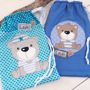 May include: Two blue drawstring bags with a sailor bear design. One bag has a turquoise background with white hearts and the other has a blue background with white dots. Both bags have the name "Levin" on them.