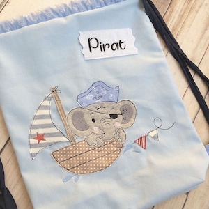 Peut inclure: Un sac à cordon bleu clair avec une étiquette blanche qui dit "Pirat". Le sac présente un motif brodé d'un éléphant gris portant un chapeau de marin bleu et un cache-œil, naviguant dans un bateau brun avec une voile rayée rouge et blanche.