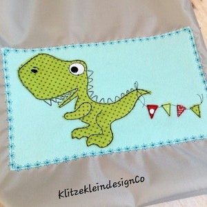 May include: A green dinosaur with white eyes and a polka dot pattern is holding a string of colorful flags. The dinosaur is on a light blue background with a blue and white stitched border. The text "KlitzekleindesignCo" is at the bottom of the image.